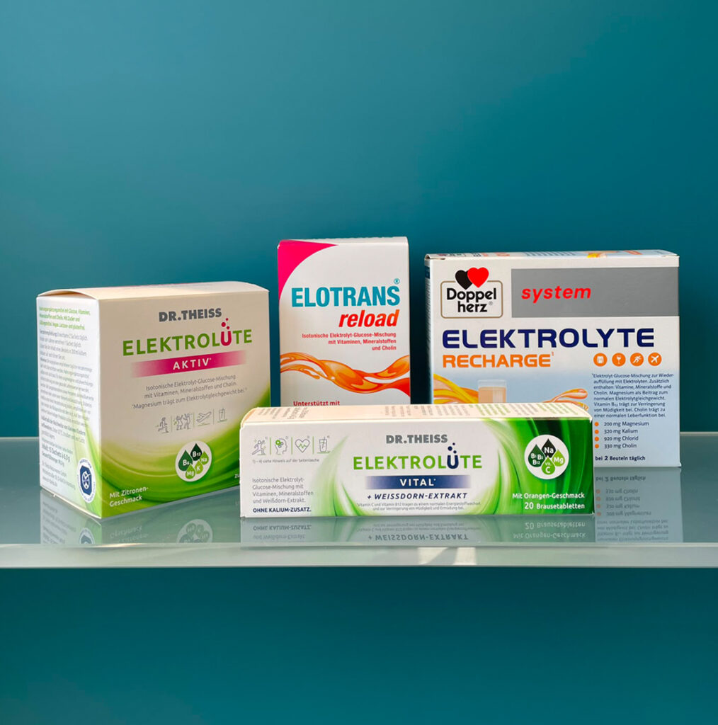 Vier verschiedene Elektrolyt-Präparate der Marken Dr. Theiss, Elotrans und Doppelherz stehen ordentlich aufgereiht auf einer spiegelnden Oberfläche vor einem blau-grünen Hintergrund.