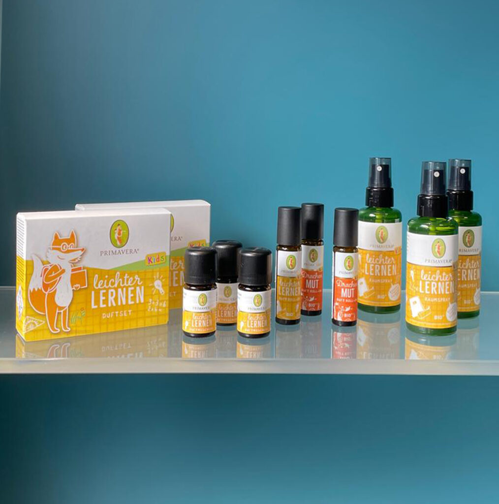 Mehrere Primavera Aroma-Roll-Ons und Raumsprays für Kinder auf einem transparenten Regal vor einer blauen Wand.