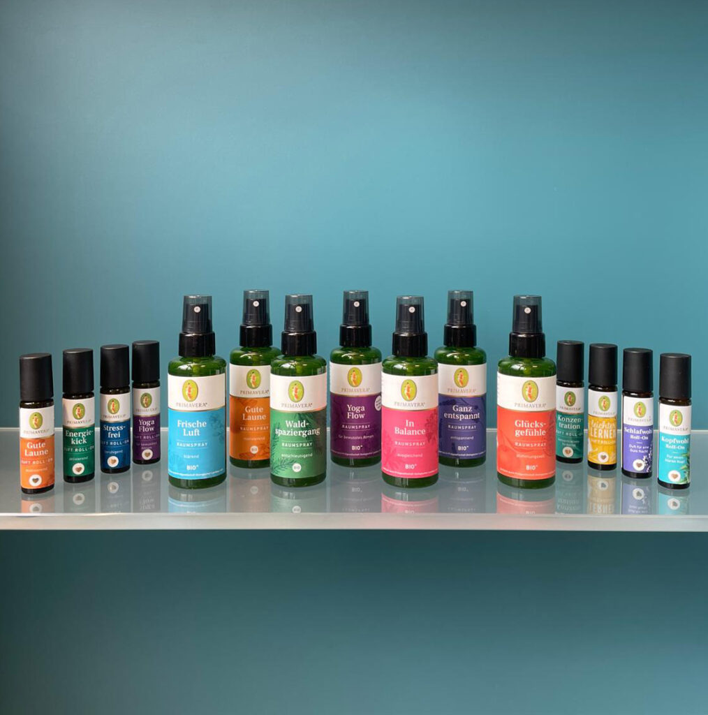 Eine Reihe verschiedener Aroma-Roll-Ons und Raumsprays der Marke Primavera steht ordentlich aufgereiht auf einem Glasregal vor einem blau-grünen Hintergrund.