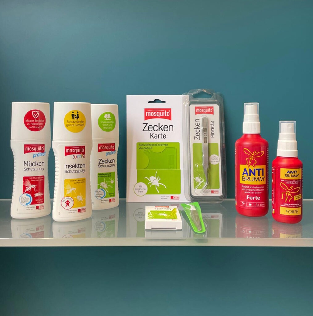 Das Bild zeigt verschiedene Mücken- und Zeckenschutzsprays sowie Werkzeuge zur Zeckenentfernung auf einem Glasregal vor blauem Hintergrund.