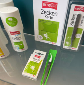 Das Bild zeigt eine Nahaufnahme von Zeckenschutz- und Entfernungsprodukten auf einer Glasfläche vor grauem Hintergrund.