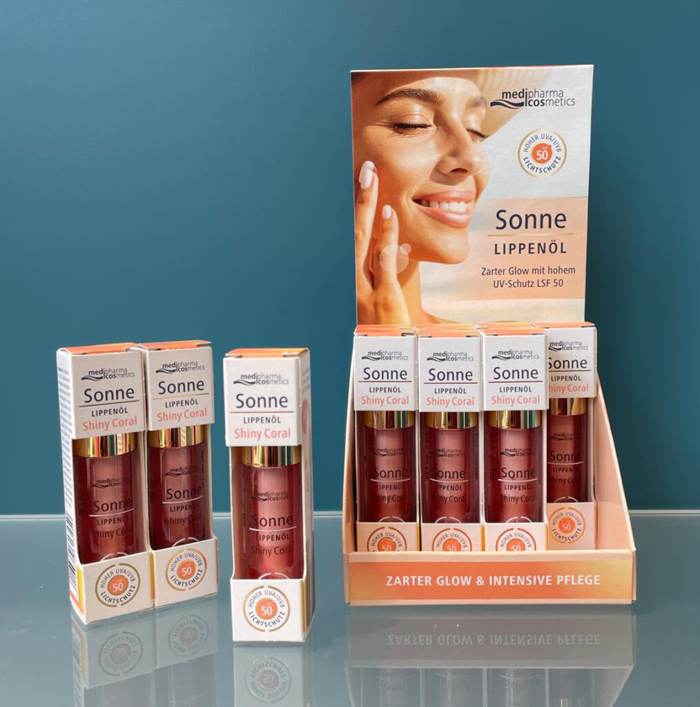 Das Produktbild zeigt mehrere Faltschachteln und einen Aufsteller des medipharma cosmetics Sonne Lippenöls in der Nuance Shiny Coral mit integriertem Lichtschutzfaktor 50 vor einem schlichten, blauen Hintergrund