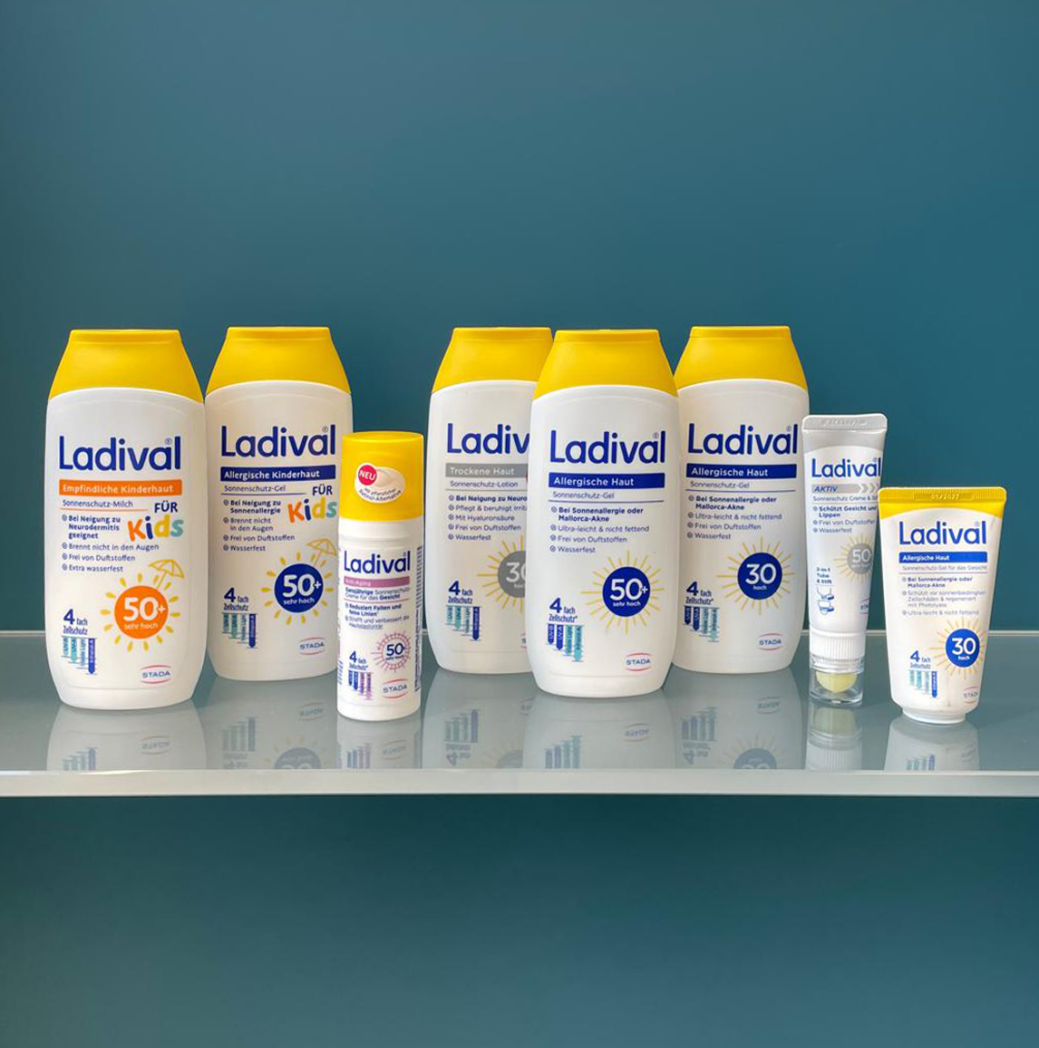 Das Produktfoto zeigt eine Auswahl an Ladival-Sonnenschutzprodukten für verschiedene Hauttypen – von Kinderhaut über allergische bis hin zu trockener Haut – aufgereiht auf einer spiegelnden Glasfläche vor blauem Hintergrund.